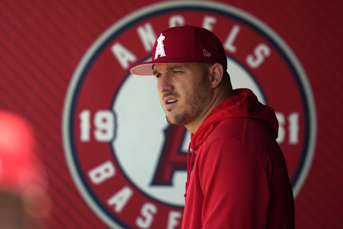 The Angels' Mike Trout stands in the dugout with a red hoodie near the team's logo