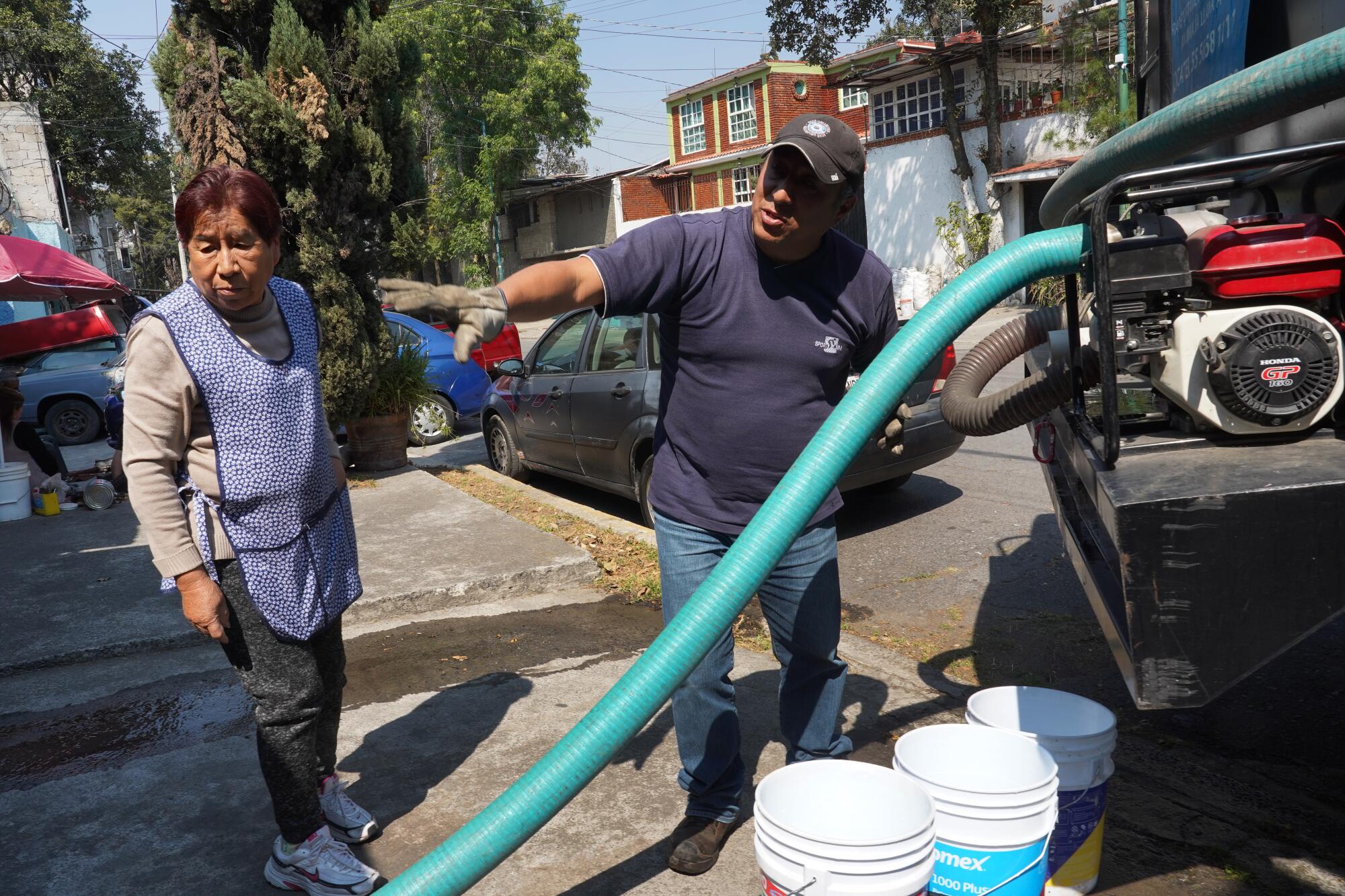 A woman stands on the sidewalk as a man holds a hose that is pumping water from a truck into a cistern at her home. 
