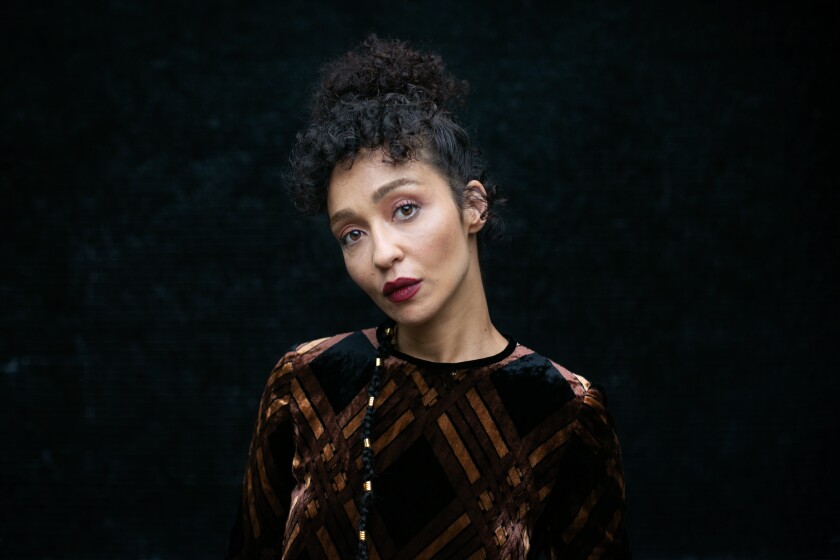 A portrait of Ruth Negga in a brown and gold striped dress against a dark background.