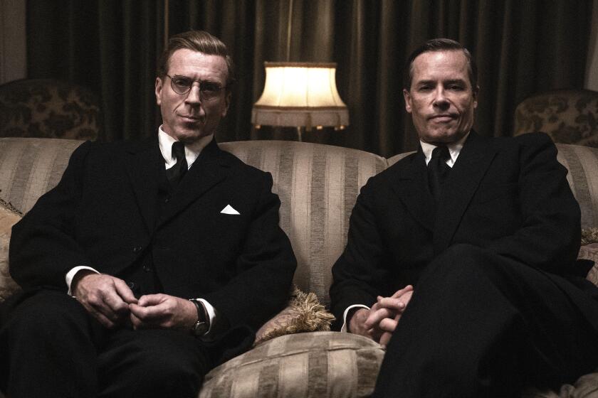 Two men in dark suits sit on a finely appointed sofa