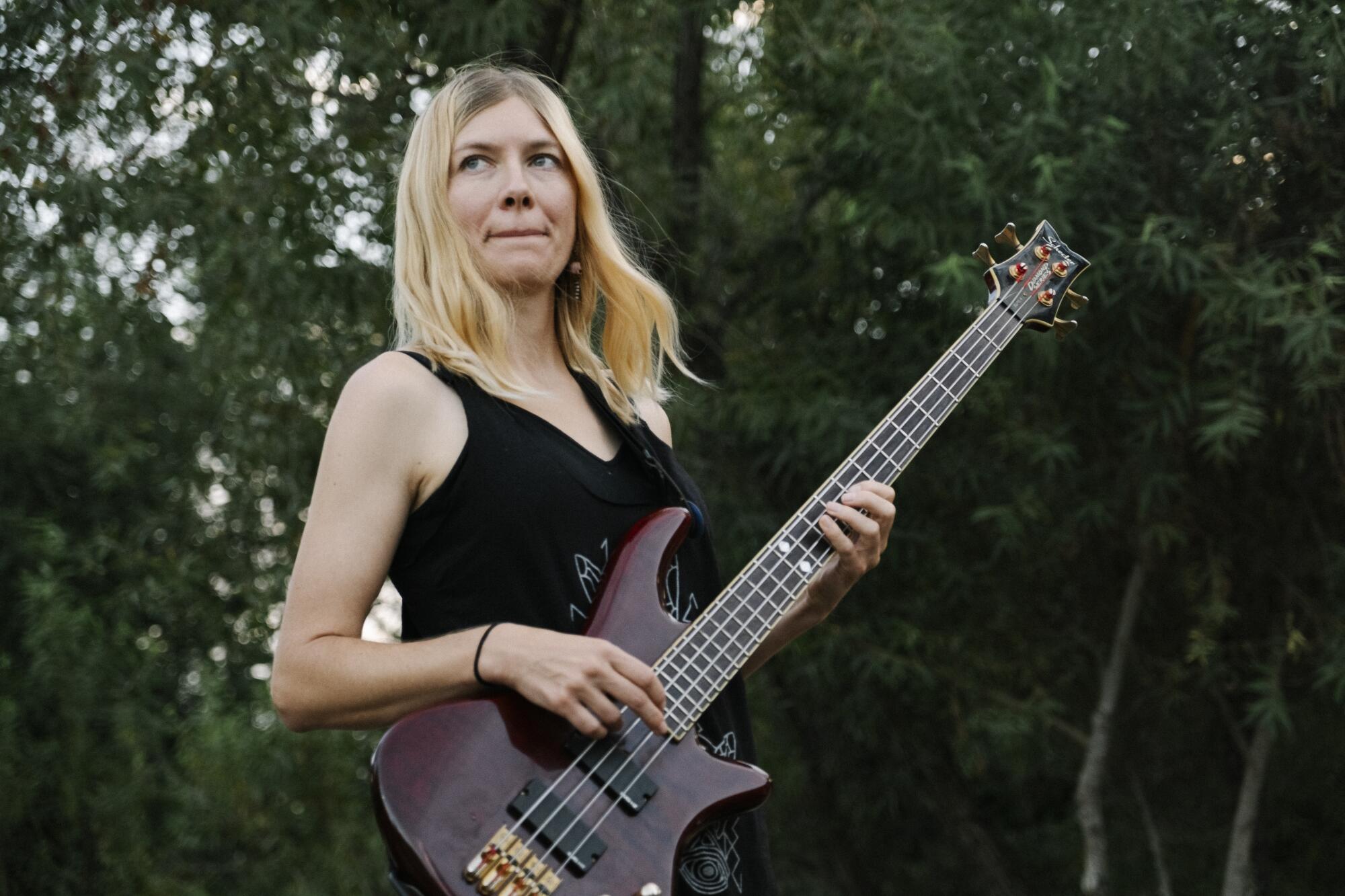 A blond woman stands outdoors holding a bass guitar.