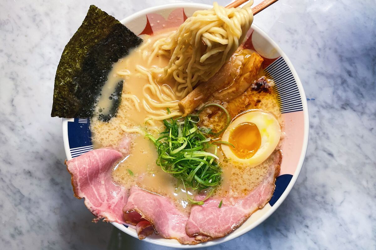 An overhead photo of ramen featuring pork, half an egg, green onions and nori. From a corner, chopsticks lift noodles.