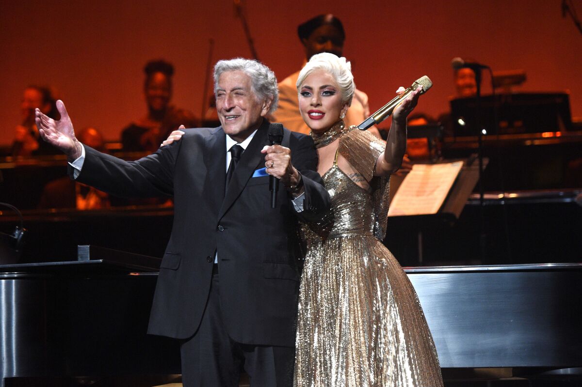 An elderly male singer and a blonde female singer acknowledge audience applause from stage.