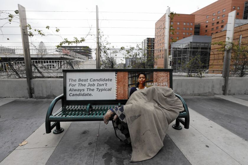 A homeless person sleeps beneath a blanket on a bus bench on South Grand Avenue.