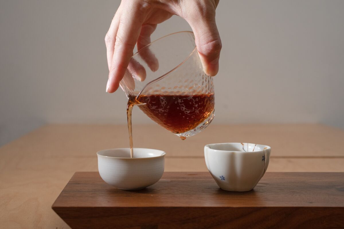 Pouring tea from a glass beaker into two white ceramic cups on a wooden board.