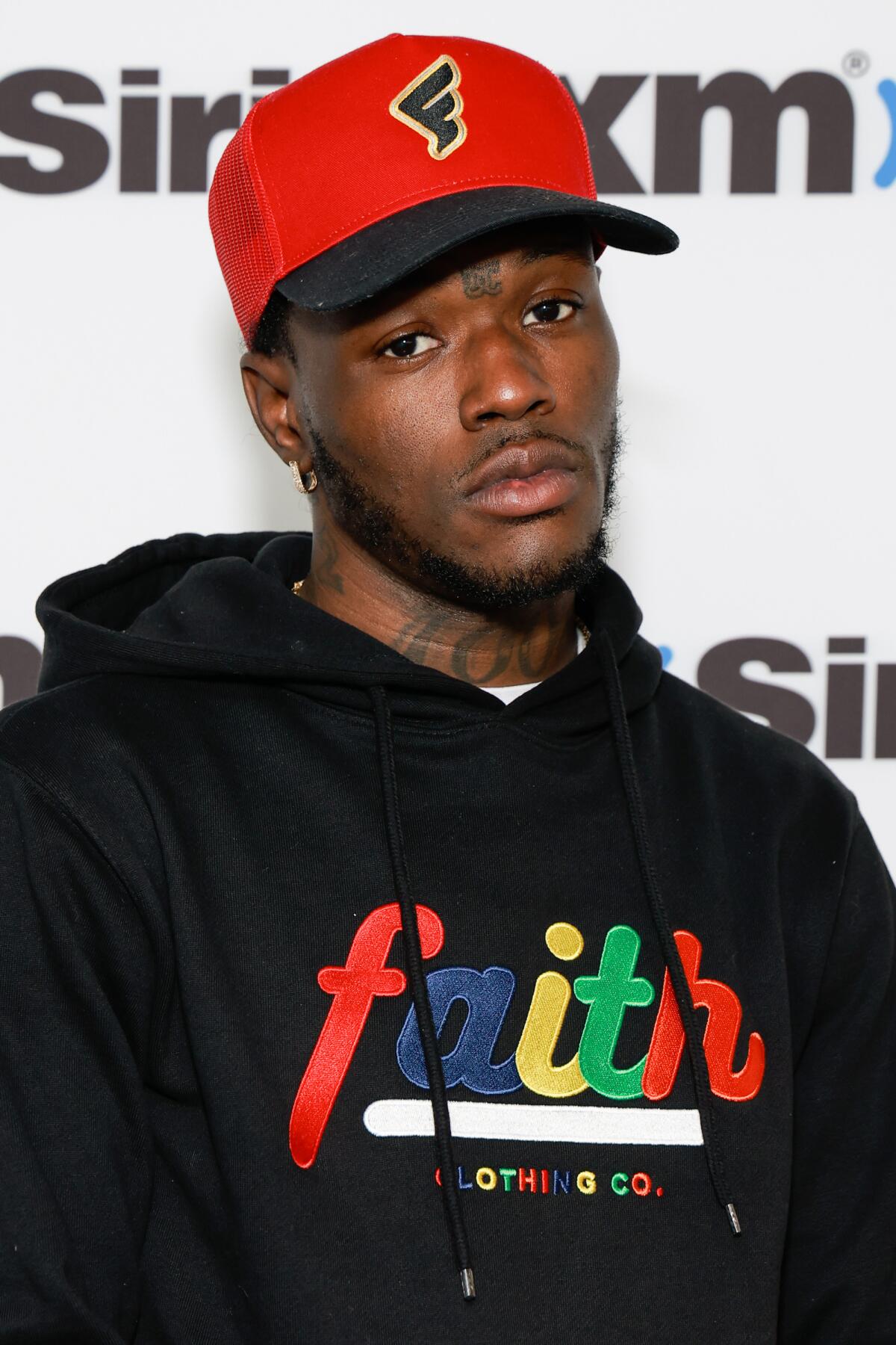 DC Young Fly is posing with a serious face while wearing a red baseball cap and a black hoodie with multicolor "faith."