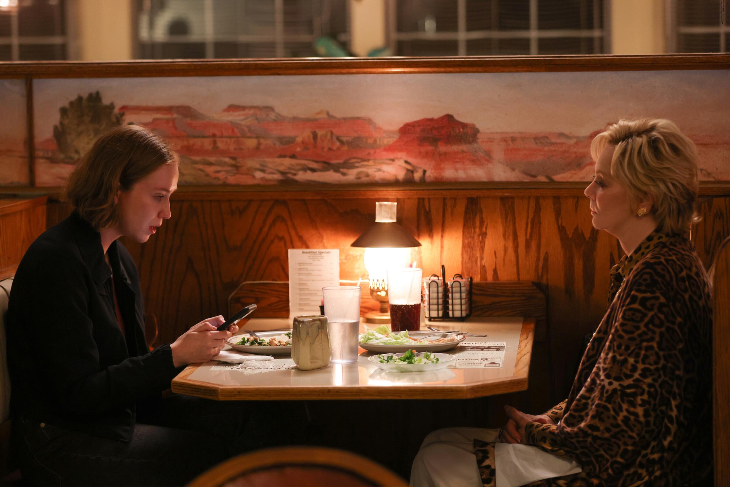 A younger woman on a phone, left, and an older woman sit across from each other at a restaurant 