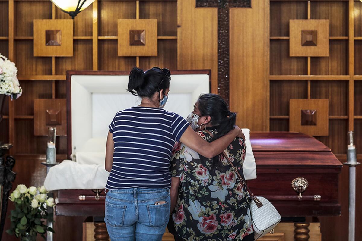 Two women stand at a casket, one with her arm around the other.