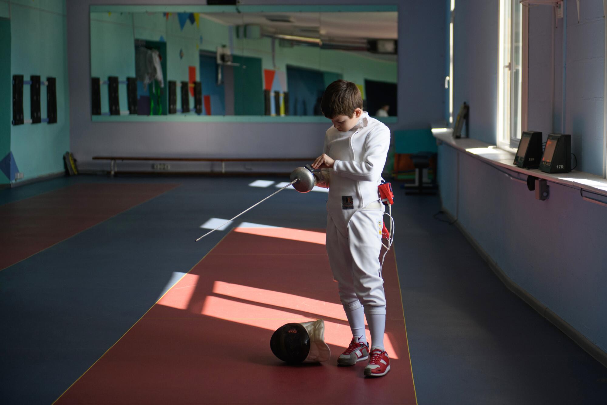 Sunlight casts stripes on the floor as a child in fencing gear gets ready to practice.