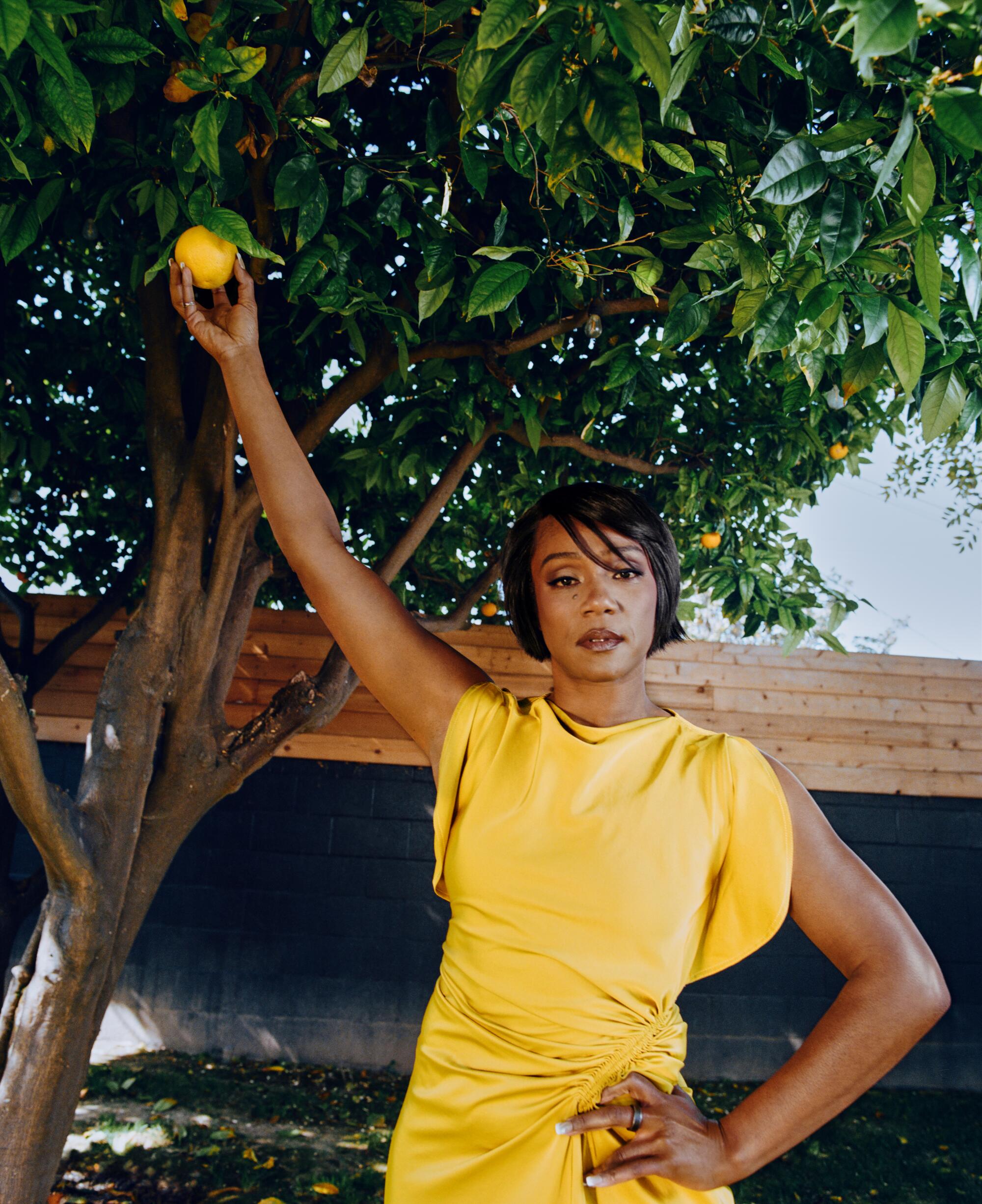 Tiffany Haddish, in a yellow dress, reaches one arm up to pick a piece of fruit off a tree at her home