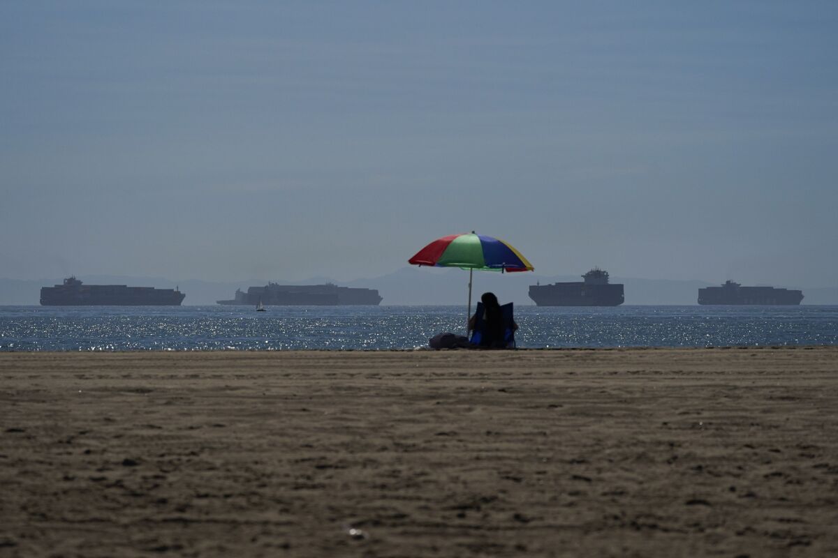 A person lounges on a beach in Seal Beach as container ships wait to dock.