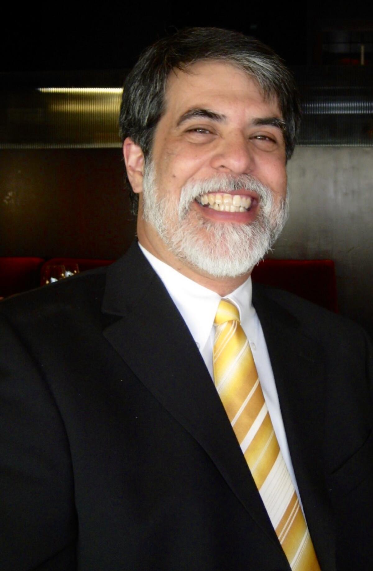 A man with black hair and white facial hair wears a black suit and yellow-stripe tie