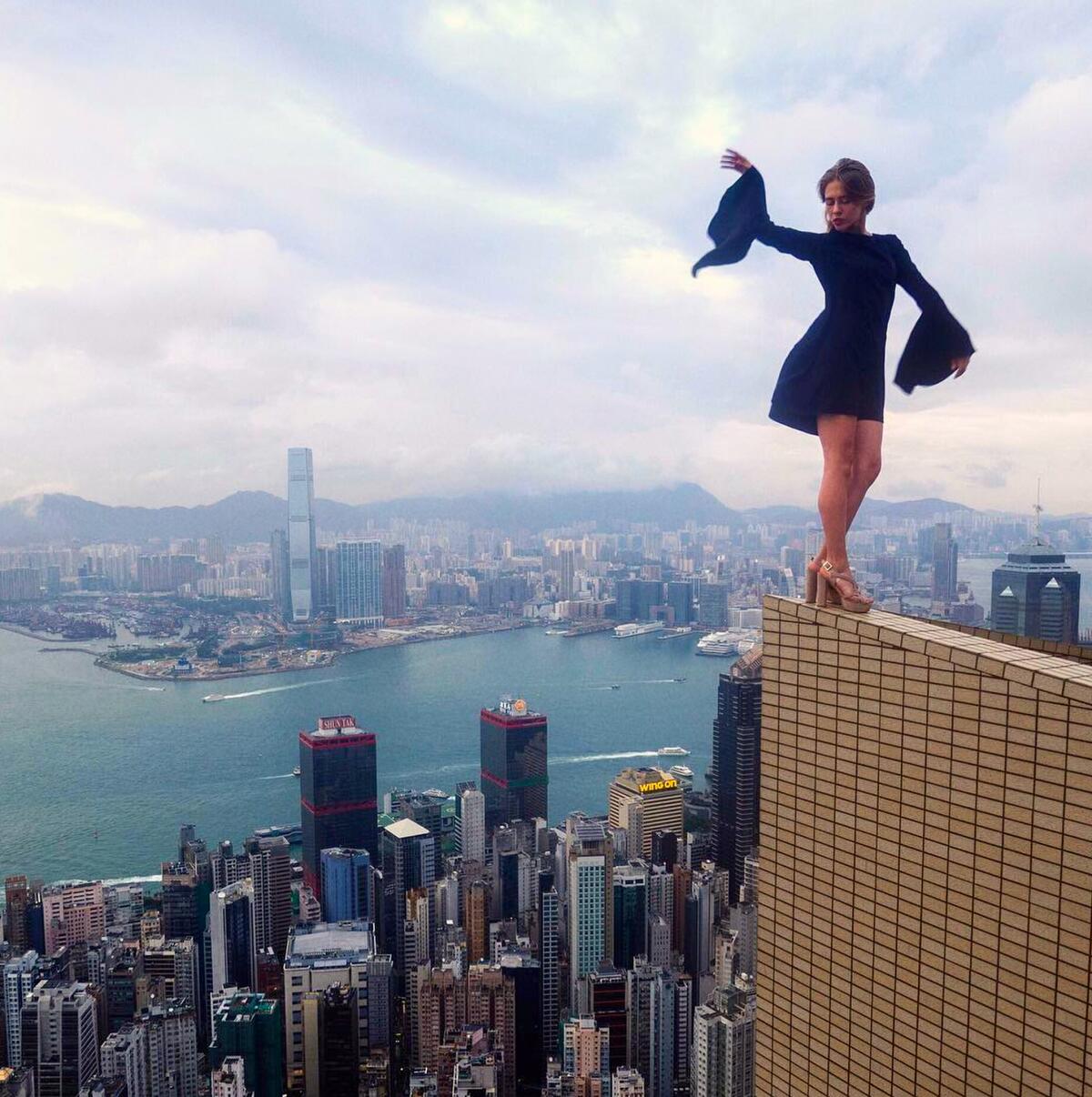 A woman in high heels and a black dress poses on the ledge of a high building.
