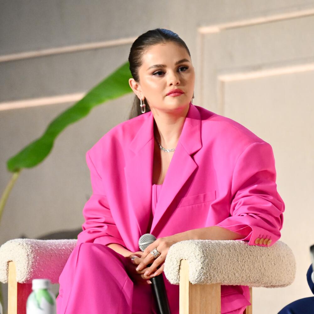 A woman in a pink suit, her hair slicked back in a ponytail, sits holding a microphone