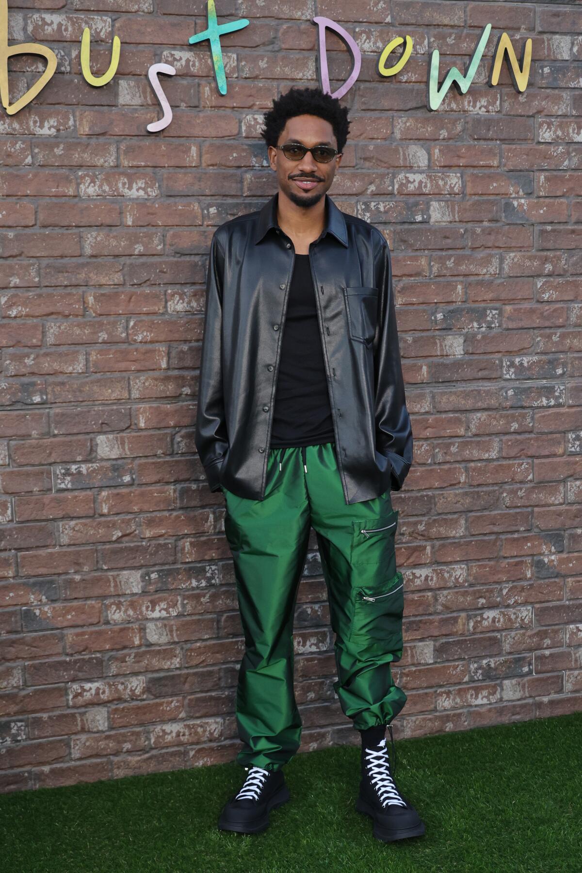 A smiling man in sunglasses, a black jacket and green pants stands in front of a brick wall.