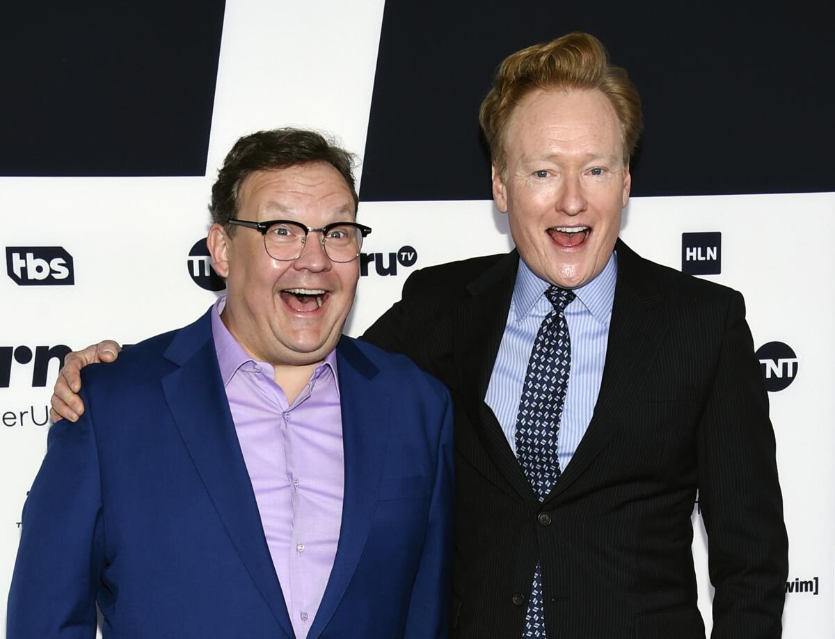 Andy Richter and Conan O'Brien pose together in suits while smiling with their mouths open.