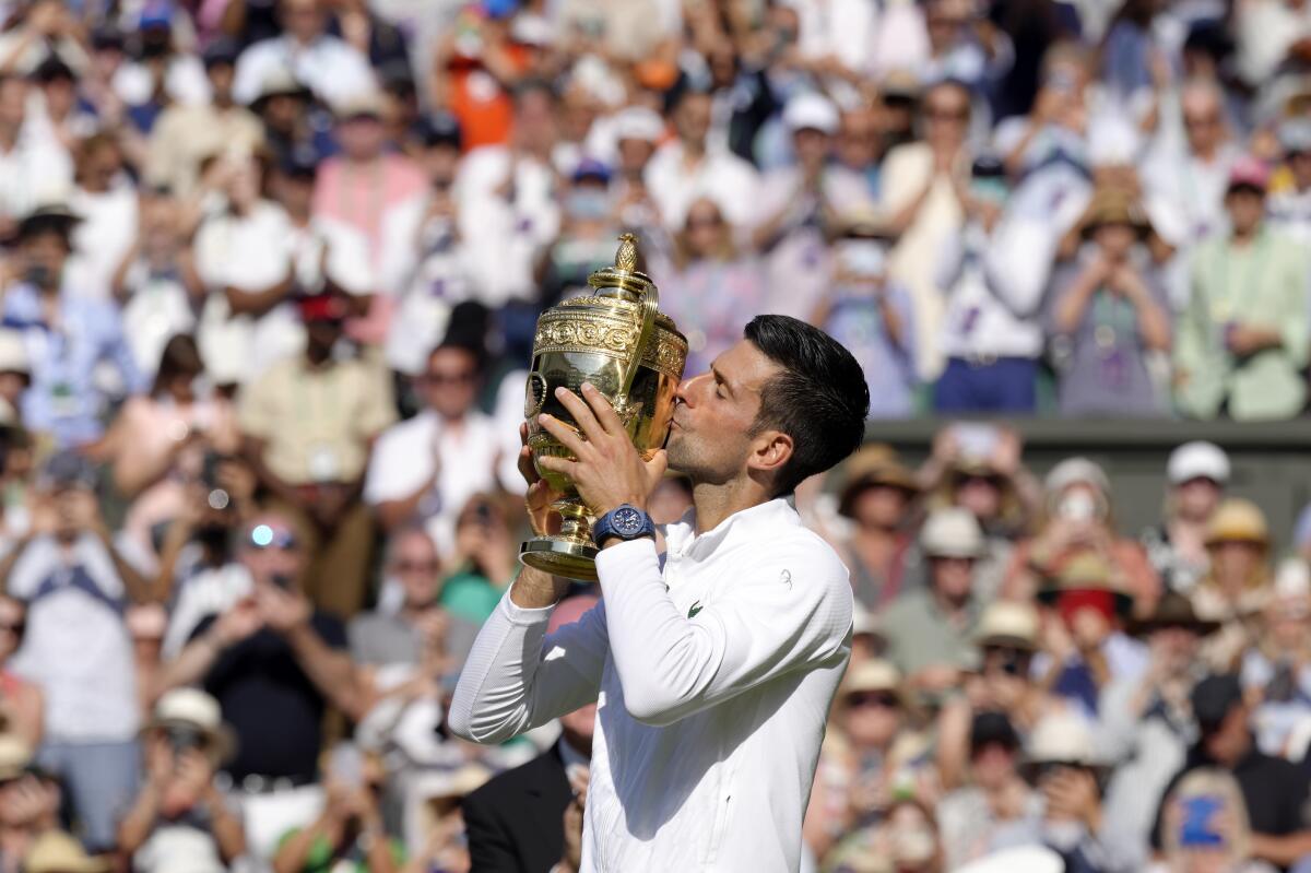 Novak Djokovic kisses the winner's trophy in front of a crowd in stands.
