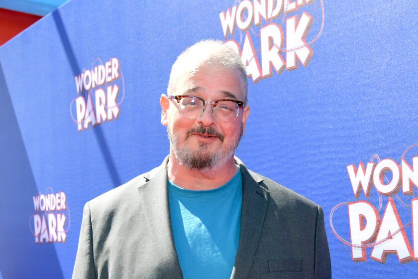 Ken Hudson smiles and poses while wearing a dark gray suit and a blue t shirt and glasses