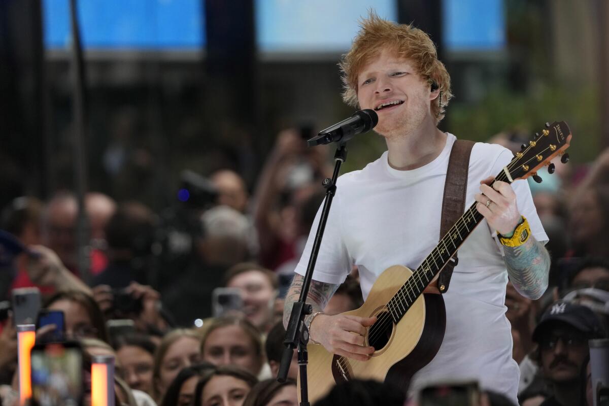 Ed Sheeran plays guitar and sings into a mic in front of numerous fans at an outdoor concert