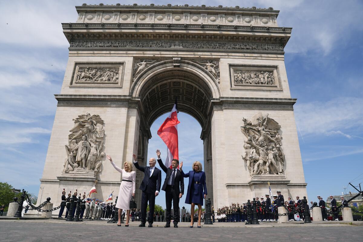 Two men flanked by two women stand and wave in front of the Arc de Triomphe in Paris.