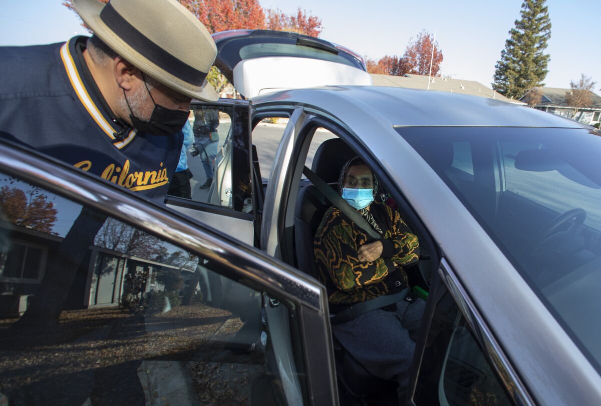 A man in a hat and mask stands at an open car door talking to a woman in the passenger seat.