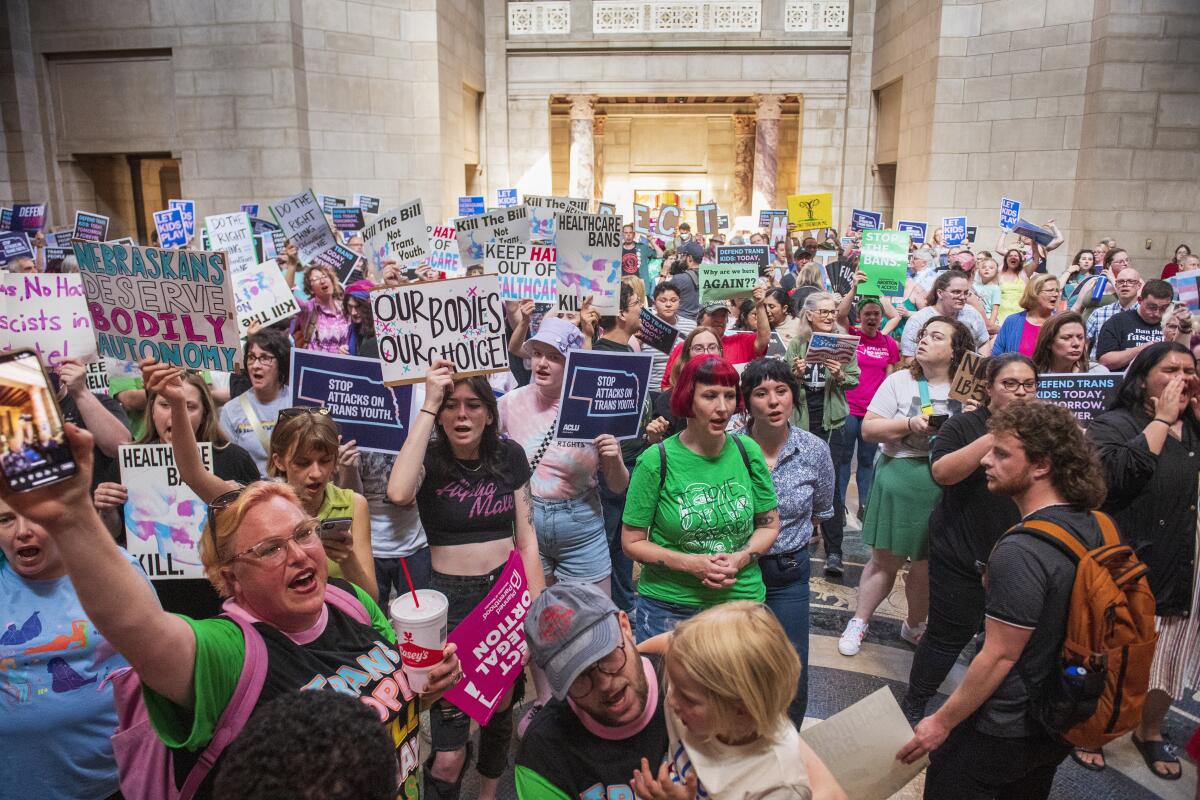 A large crowd of people standing in a room and raising signs with messages including "Nebraskans deserve bodily autonomy"