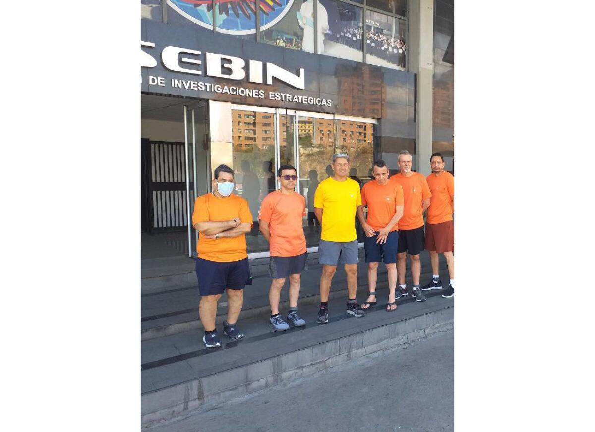 Six men in orange and yellow T-shirts and shorts stand outside.