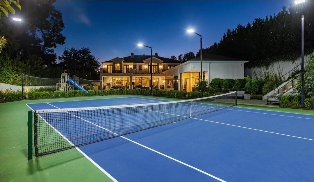 The blue tennis court at night with the house and trees in the background.