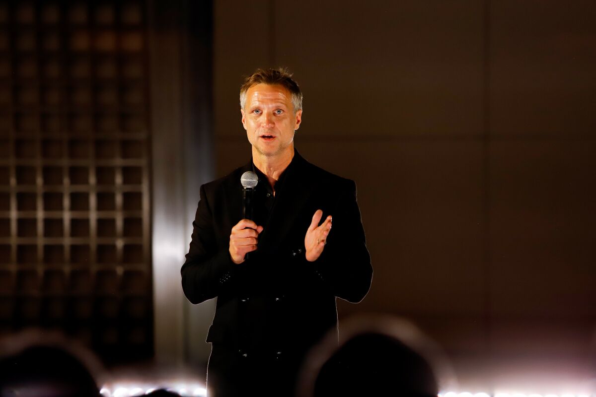 A man in a dark suit speaks into a microphone before a seated audience.