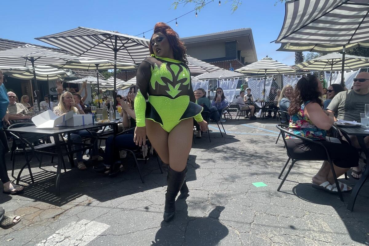 A drag queen in a black and fluorescent yellow leotard struts through a dining area set up on a parking lot