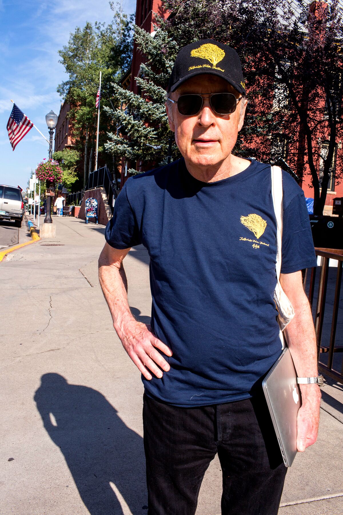 A man stands outside in the sun wearing a baseball hat, dark glasses and dark clothing.