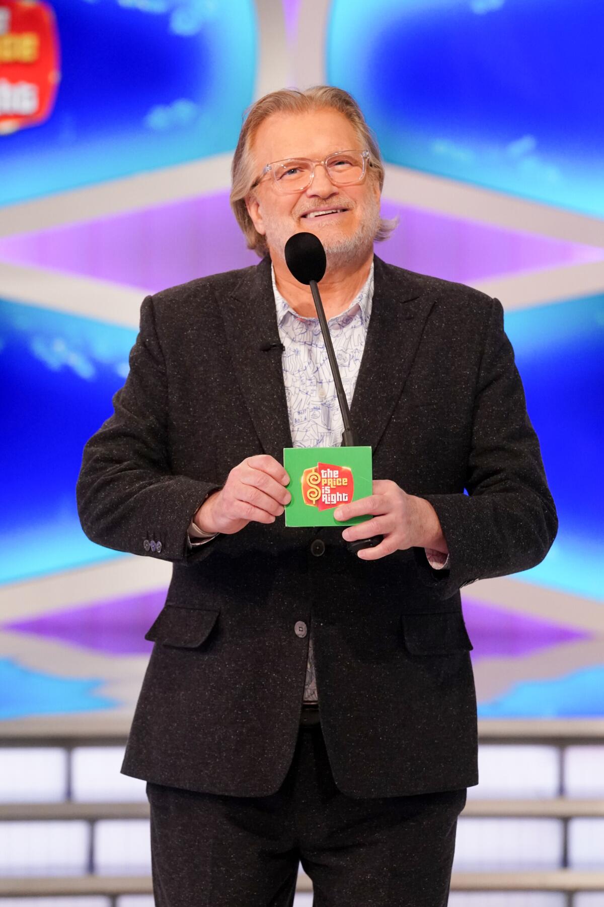 Drew Carey wears a black suit and smiles in front of a microphone while holding a green cue card.