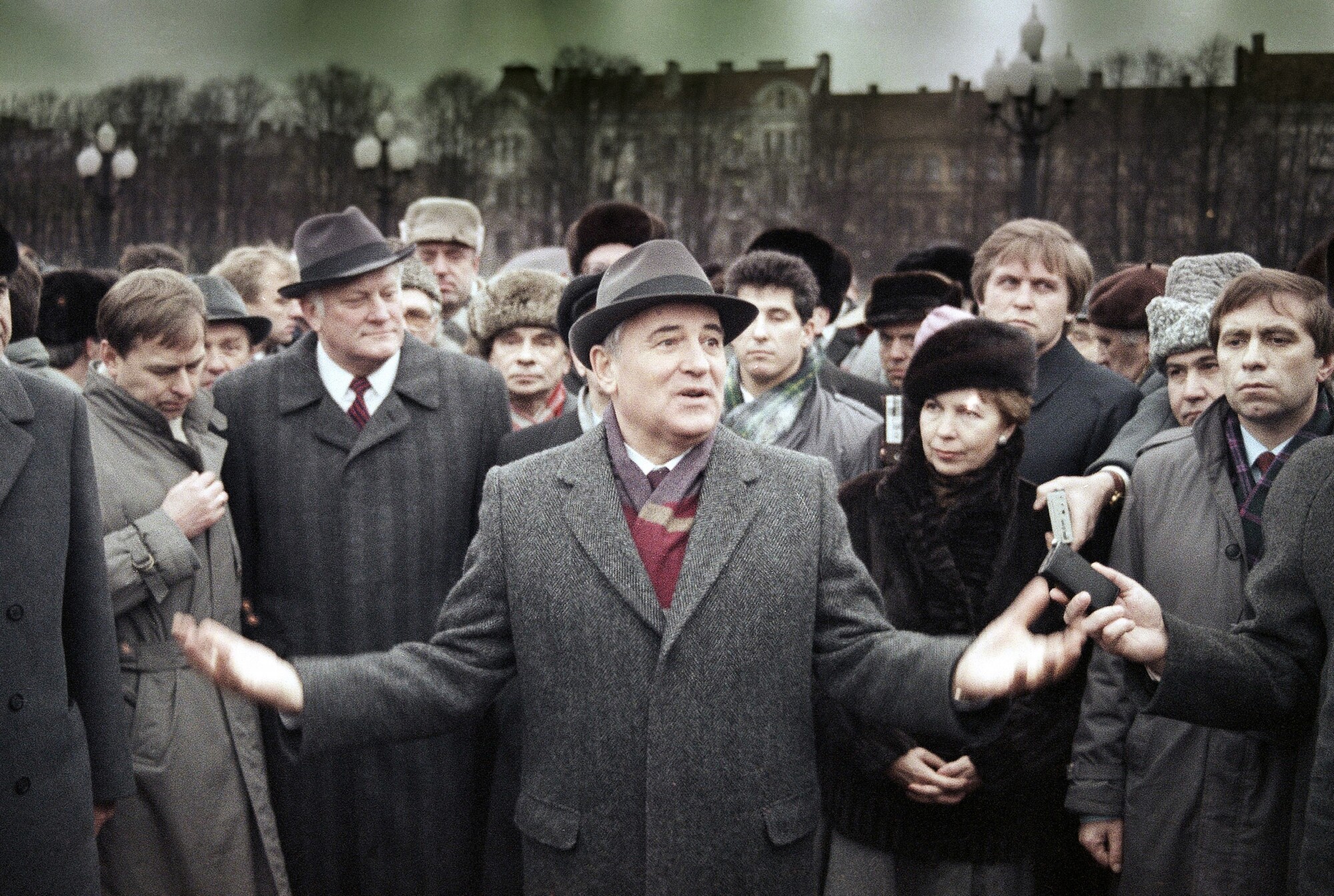Gri paltolu ve şapkalı bir adam, ellerini avuçları yukarıya kaldırmış, etrafını saran bir kalabalığa konuşuyor. 