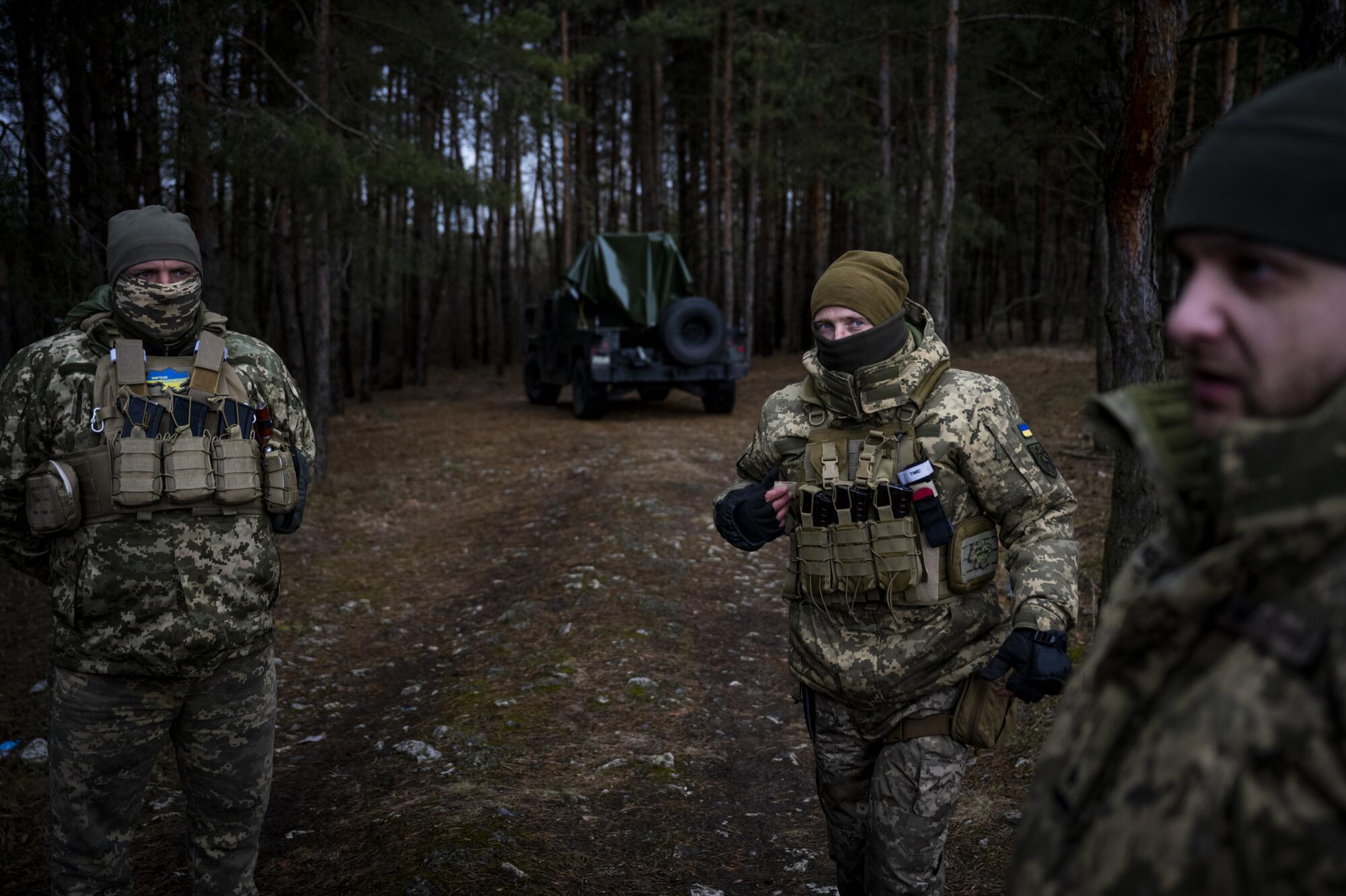 Troops in camouflage fatigues, beanies and balaclavas standing in a forest with military equipment in the background