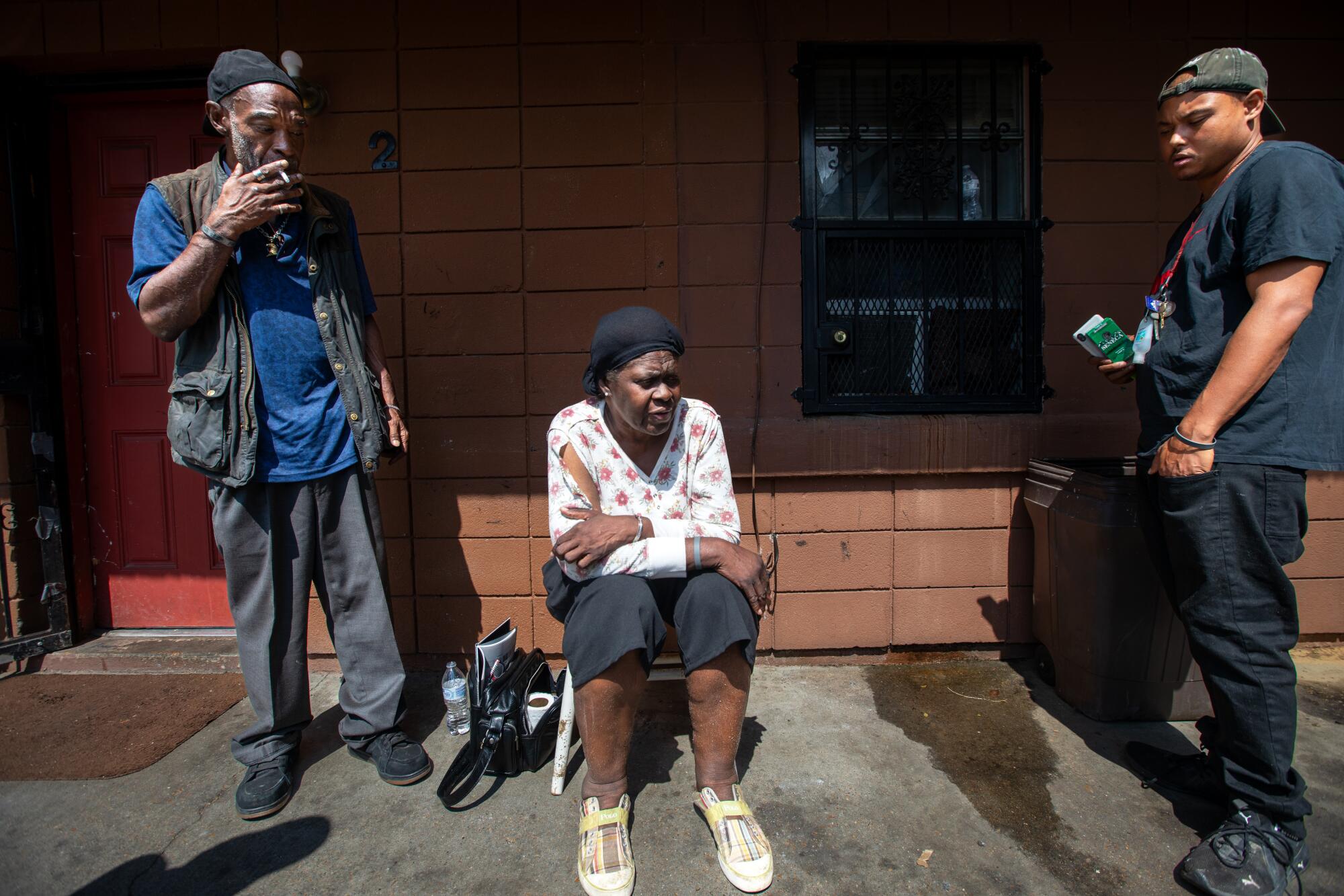 A woman sits outside a building, flanked by two men.