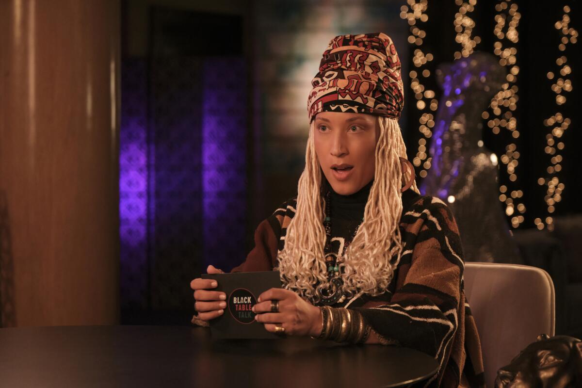 Robin Thede is dressed as character with blonde braids who is wearing a headwrap and long-sleeve top. 