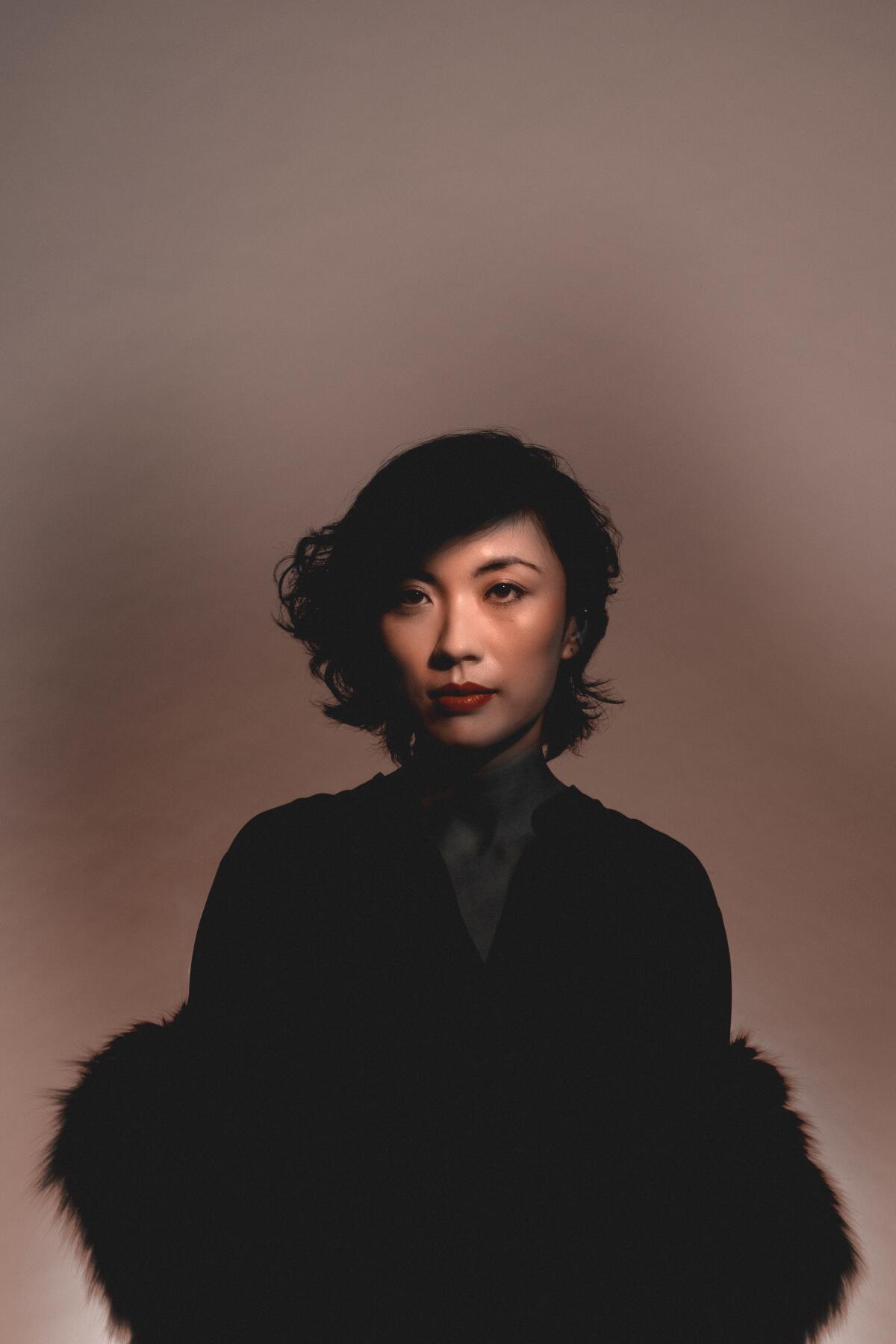 A woman in a feathery black dress poses sternly against a golden-tan background.