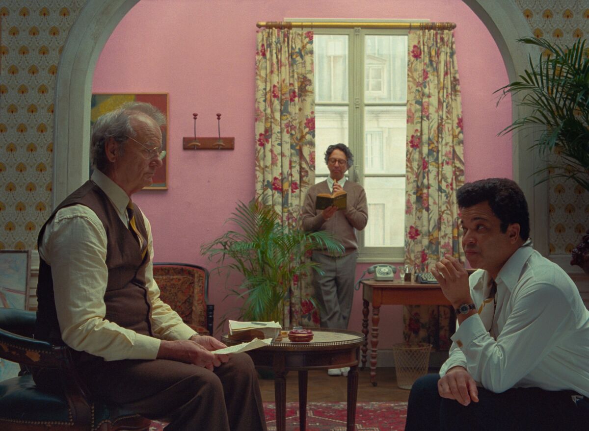 Two men sit facing each other, while another stands at a window reading a book in the film "The French Dispatch."