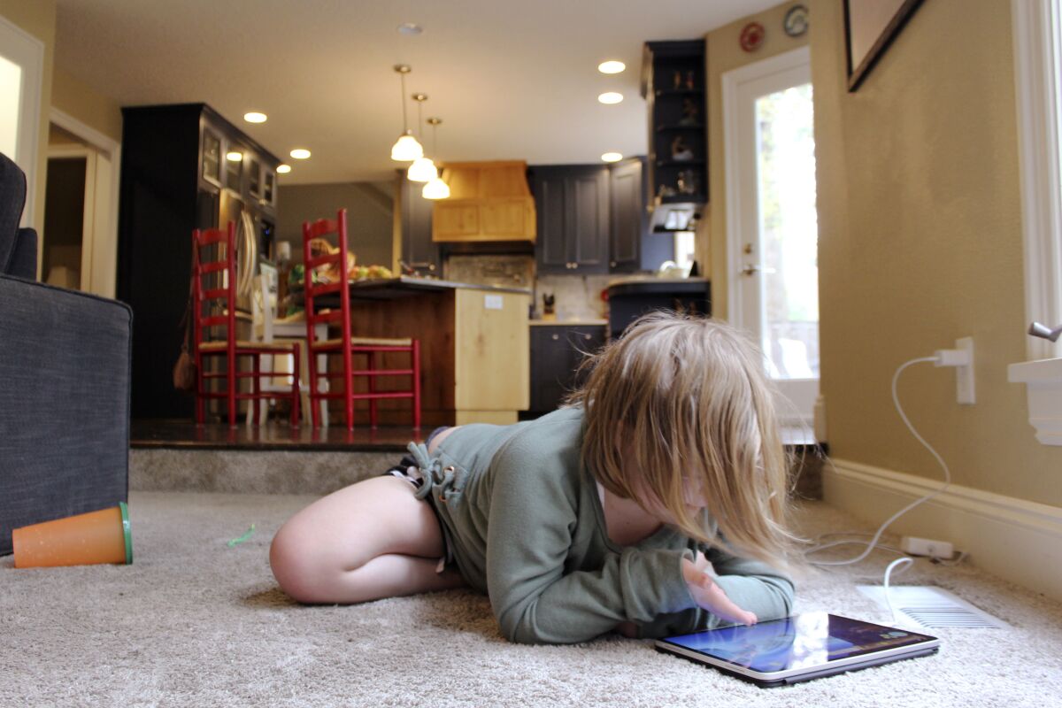 Lizzie Dale sprawls on the floor to play games on an iPad as her siblings work on school work in the kitchen behind her in their home in Lake Oswego, Ore., Oct. 30, 2020. In Oregon, one of only a handful of states that has required a partial or statewide closure of schools in the midst of the COVID-19 pandemic, parents in favor of their children returning to in-person learning have voiced their concerns and grievances using social media, petitions, letters to state officials, emotional testimonies at virtual school board meetings and on the steps of the state's Capitol. (AP Photo/Sara Cline)