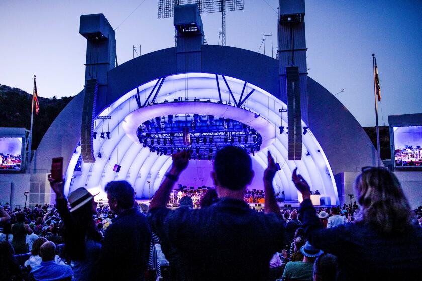Silhouettes of concert attendees raising their hands in front of the Hollywood Bowl, which has a purple hue