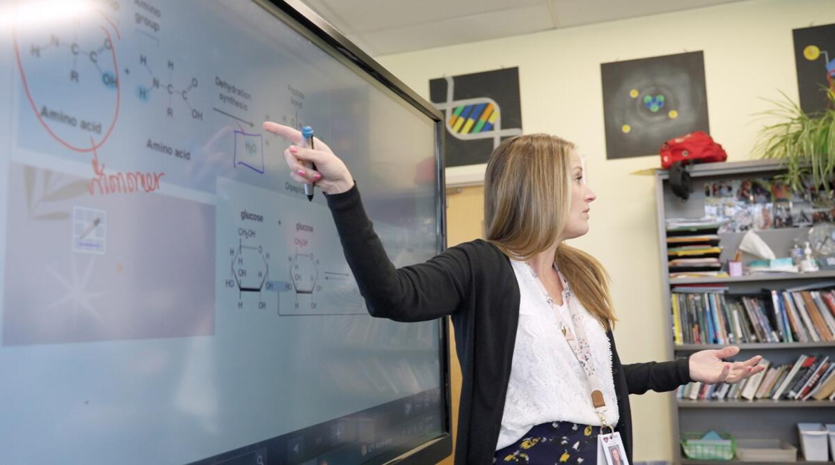 A high school chemistry teachers points to writings on a white board.