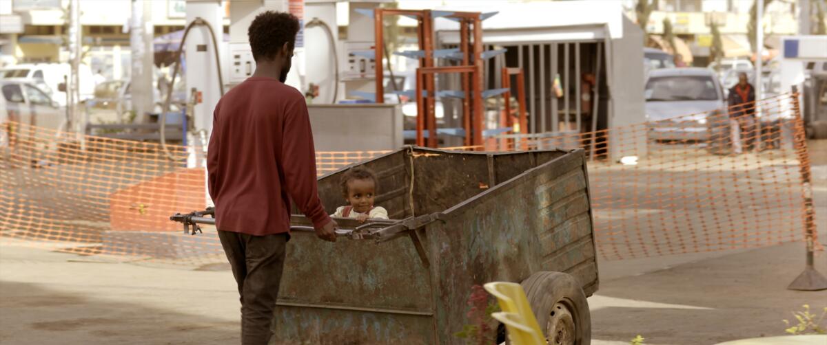 A man pushes a small child in a cart in the movie "Running Against the Wind."