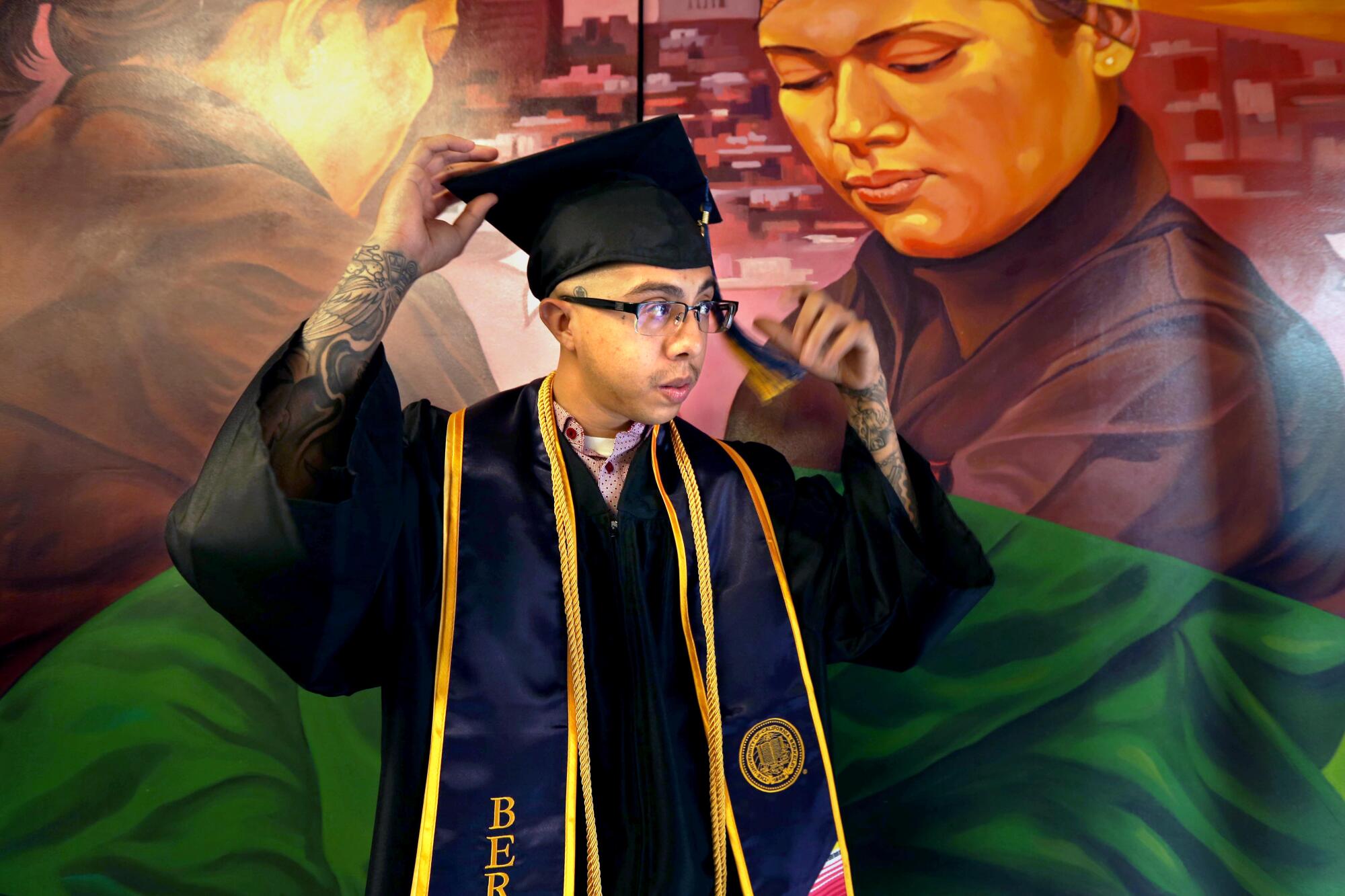 A student standing in front of a large mural adjusts his graduation cap.