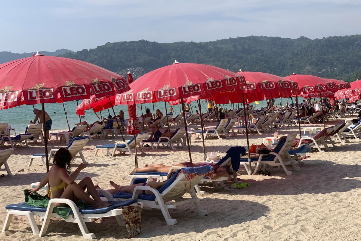 Tourists lounge under umbrellas on the beach with water and green hills in the distance.
