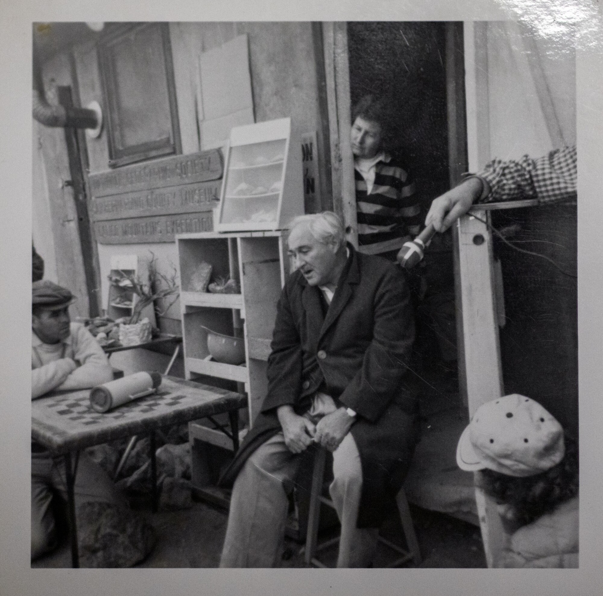 An older man sits on a stool and speaks while another person holds a microphone near his face