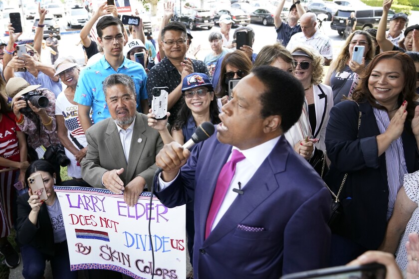 Larry Elder speaks into a microphone surrounded by supporters