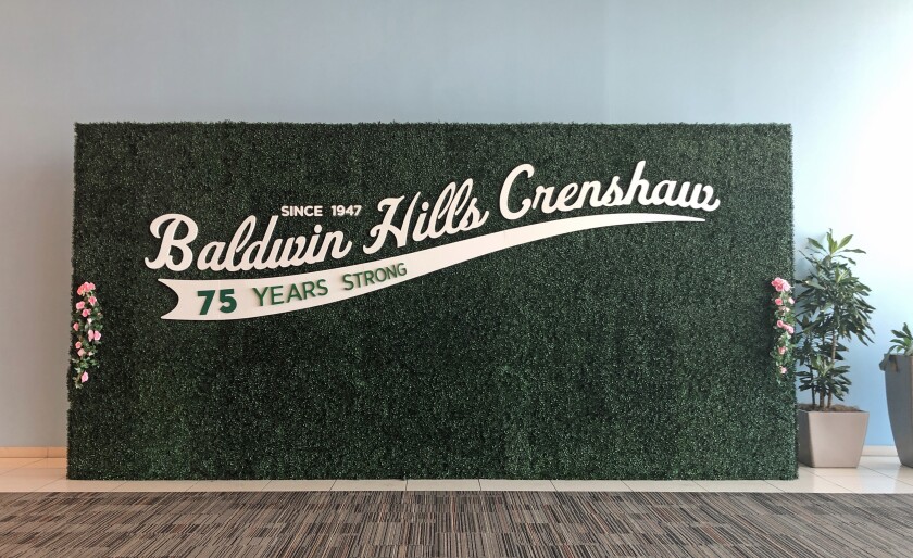 A green backdrop features a sign in white cursive that reads: "Baldwin Hills Crenshaw: 75 Years Strong"