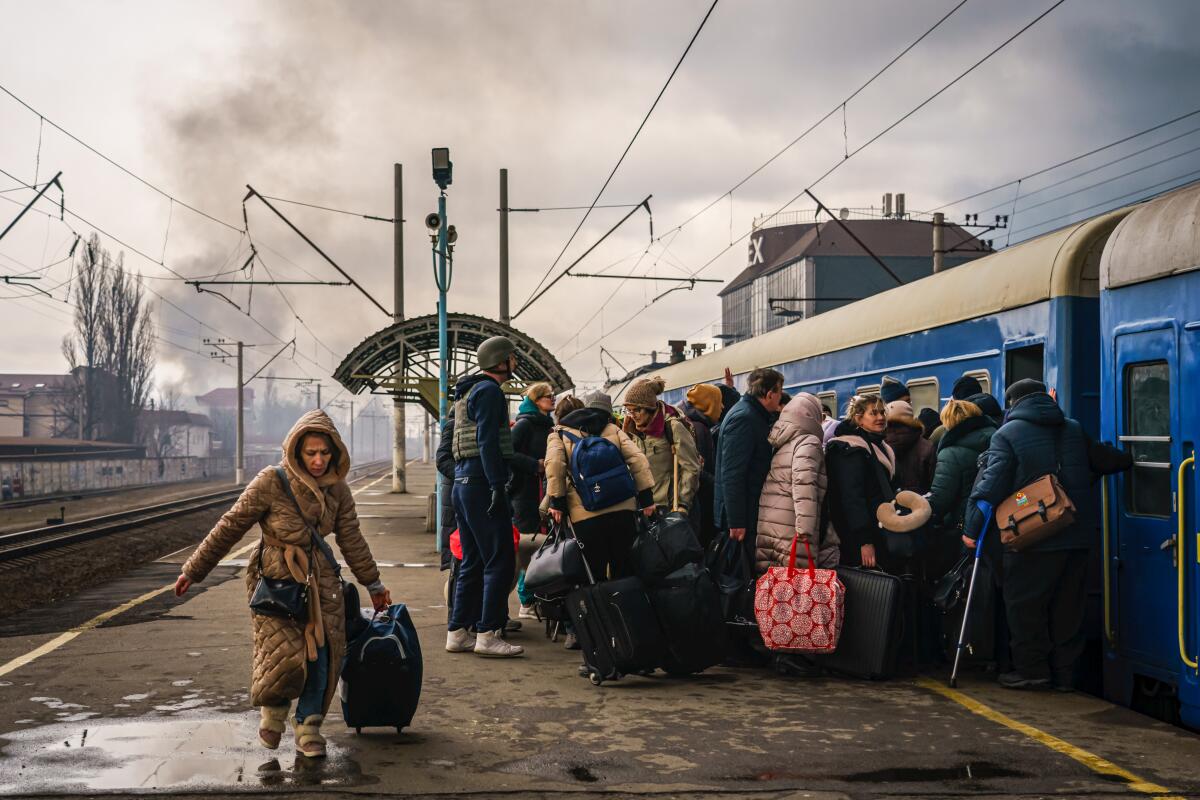 Civilians dressed for winter line up to board a train as a woman walks away, pulling her luggage