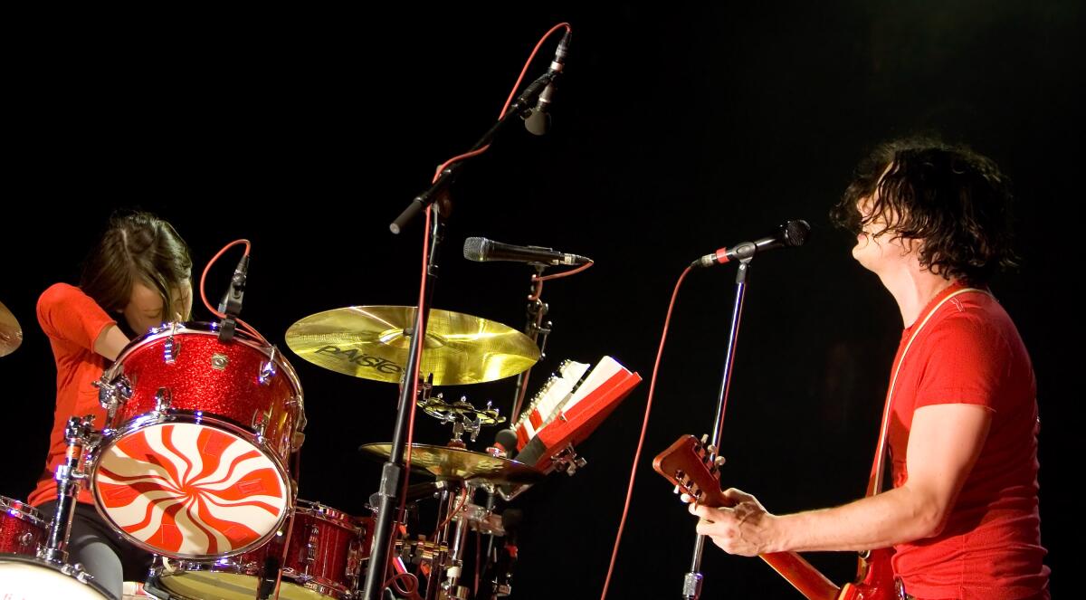 A woman in red playing a red drum set and a man in red playing guitar and singing into a microphone