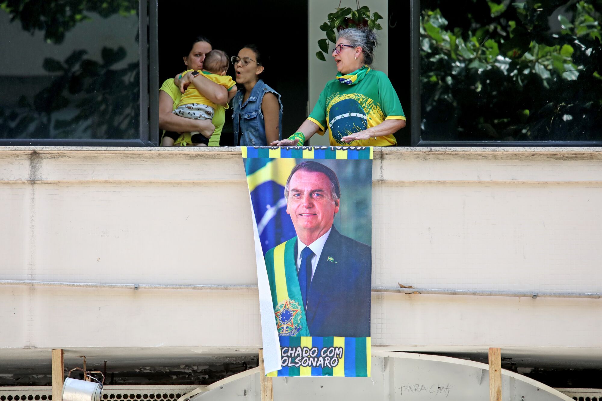 Three women, one of whom is holding a baby, stand behind a poster of a man in suit and tie wearing a green-and-yellow sash 